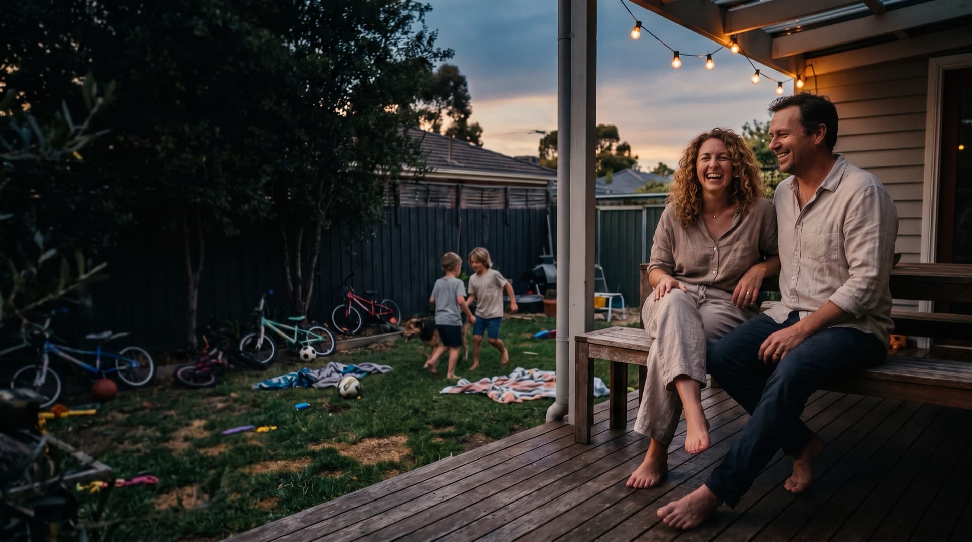 A family unwinding at home at dusk, parents relaxed on the back deck while the kids play on the lawn below