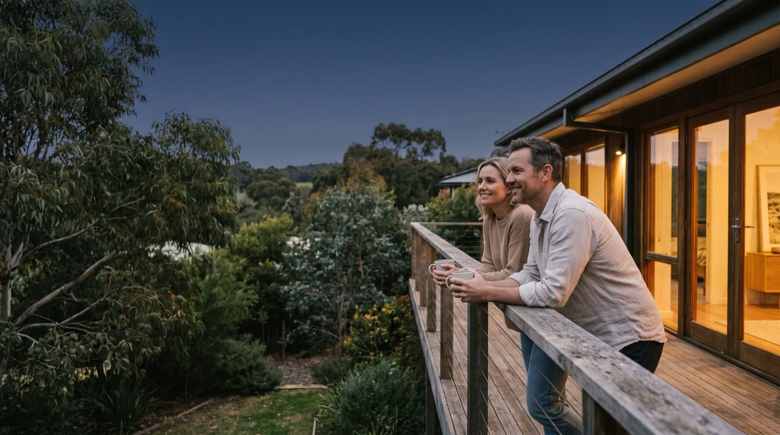 A couple on their deck at dusk with warm light from the house behind them, looking out over the garden